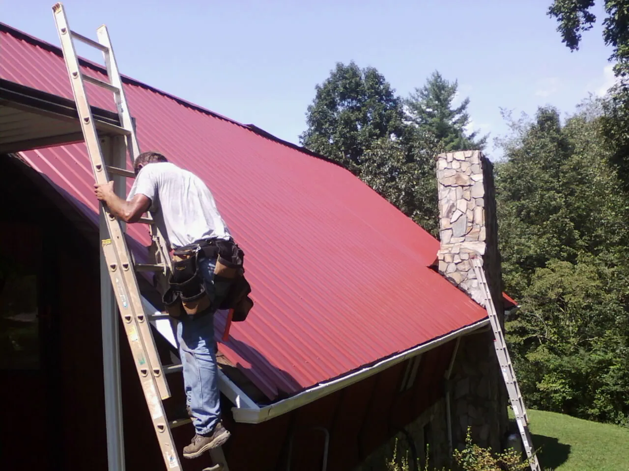 Roofer on a ladder beside a red metal roof and stone chimney — Soffit & Fascia Repair by Roofing Brothers in Plaistow
